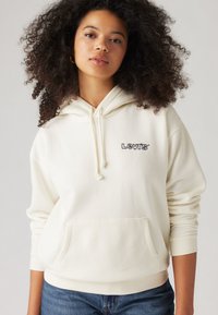 Ung kvinna med lockigt hår iklädd en krämfärgad hoodie från Levi's och blå jeans mot en enkel ljus bakgrund.