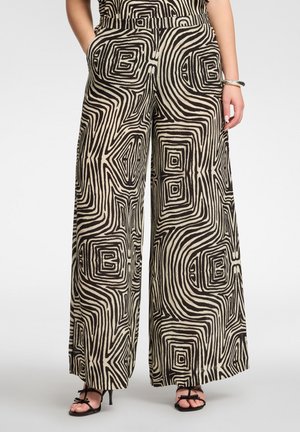 Kobieta nosząca spodnie z szerokimi nogawkami, z czarno-beżowymi abstrakcyjnymi geometrycznymi wzorami w spiralę oraz czarne sandały na wysokim obcasie z paseczkami.