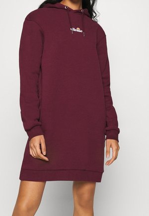 Femme portant une longue robe à capuche bordeaux avec un petit logo blanc ellesse sur la poitrine et des cordons à la capuche.