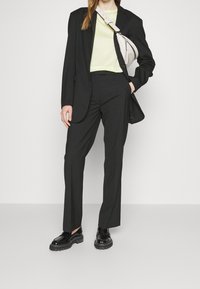Černý elegantní oblek se slim-fit kalhotami, doplněný světle zeleným topem. Obsahuje strukturované sako a lesklé černé boty. Malá bílá kabelka.