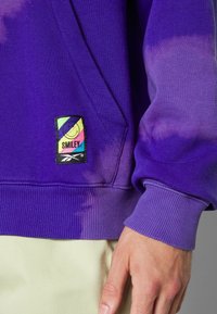 Lilanhuppari, joka on valmistettu puuvillasekoitteesta, jossa on sitruunavärisiä aksentteja ja Smiley-logopainatus edessä. Ribbaatut hihansuut ja helma tarjoavat rakennetta.