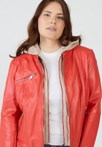 Röd läderjacka med beige huva, dragkedja framtill och sidofickor. Har en slät yta och horisontella kviltade detaljer på axlarna.