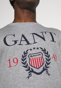 Camiseta de algodón gris con logo bordado en azul marino y rojo, en la parte izquierda del pecho, con diseño de laurel y el texto "GANT 1942" y "19".