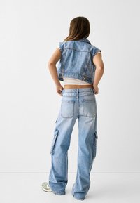 Světle modrá denimová vesta a cargo kalhoty, které mají zkrácený střih, klopové kapsy a volný střih. Hladká textura, ležérní styl.