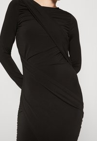 Černé šaty s dlouhým rukávem, která mají shromážděnou texturu a diagonální řasení, vyrobené z elastického materiálu, který se přizpůsobuje postavě.