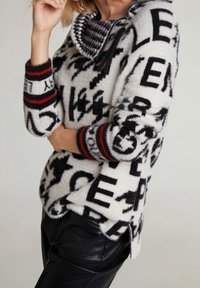 Pull surdimensionné et duveteux couleur crème avec de grosses lettres noires. Il comporte des poignets rayés noirs et rouges ainsi qu'une capuche à motif pied-de-poule.