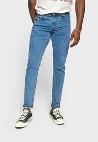 Siniset farkut slim fit -mallissa, joissa on viisi taskua, ommeltu yksityiskohta ja nilkkaan ulottuva helma, yhdistetty mustiin lenkkitossuihin.