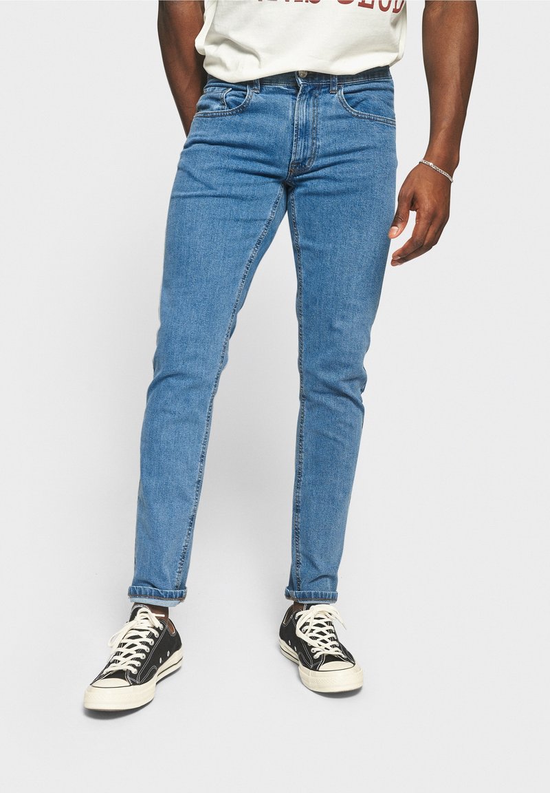 Siniset farkut slim fit -mallissa, joissa on viisi taskua, ommeltu yksityiskohta ja nilkkaan ulottuva helma, yhdistetty mustiin lenkkitossuihin.