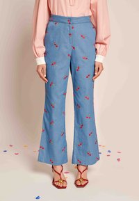 Modre kavbojke s širokimi hlačnicami in rdečimi vezeninami češenj, visokega pasu z gladko teksturo, v kombinaciji z rdečimi sandalnimi čevlji z trakovi, ki imajo zlate dodatke.