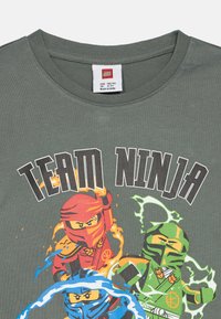 Sötétzöld pamut póló, amelyen a "Team Ninja" grafika látható, három nindzsával piros, kék és zöld színekben, elemi hatásokkal körülvéve.