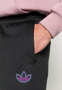 Svarta sweatpants med en sidficka. Har en flerfärgad broderad logotyp i lila och blått. Midjan är elastisk.