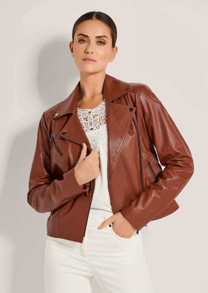 Vrouw met donker haar in een naar achteren gestylede look, die een bruine leren jas draagt over een witte kanten top en witte broek, poserend tegen een effen achtergrond.