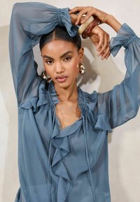 Sheer, ruffled blouse in a muted blue tone, featuring elasticized sleeves, a deep V-neckline, and a lightweight, flowing texture.