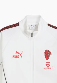 Hvid zip-up jakke lavet af glat stof. Har røde detaljer, Puma-logo, "KING" tekst og et drageemblem. Sorte sidelommer med mønstre.