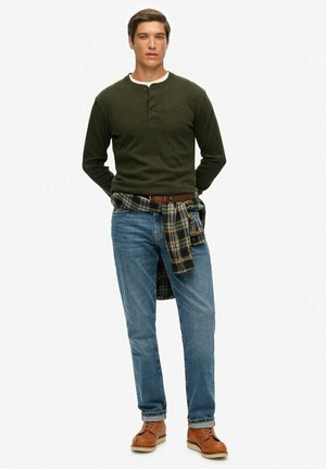 Hombre vestido con una camiseta de manga larga verde oscuro, jeans azules, cinturón marrón y una camisa de cuadros atada a la cintura, de pie.