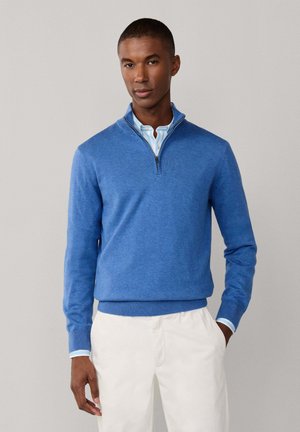 Hombre con un suéter azul de media cremallera sobre una camisa clara a rayas y pantalones blancos, de pie con una mano en el bolsillo contra un fondo gris.