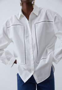 Witte blouse van katoen, met zwarte gestreepte accenten langs de borstzakken en manchetten. Losse pasvorm, lange mouwen en een ronde zoom.