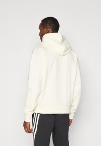 Sweatshirt branco texturizado com gola alta e capuz, apresentando punhos e bainha canelados, combinado com calças de treino cinza escuro com três riscas.