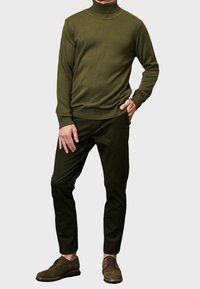 Pull en col roulé vert, avec des poignets et un ourlet côtelés, associé à un pantalon slim-fit vert foncé. Chaussures à lacets marron avec des détails de perforation subtils.