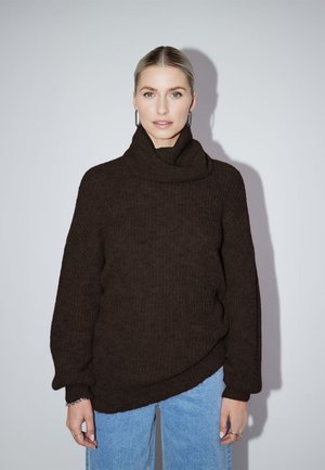 Vrouw kijkt naar voren, draagt een donkerbruine coltrui en lichtblauwe jeans, staand tegen een effen lichte achtergrond.