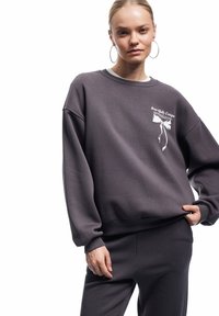 Szara bluza z miękkiego materiału, z białym nadrukiem graficznym i tekstem. Luźny krój z ściągaczami przy mankietach i pasie.