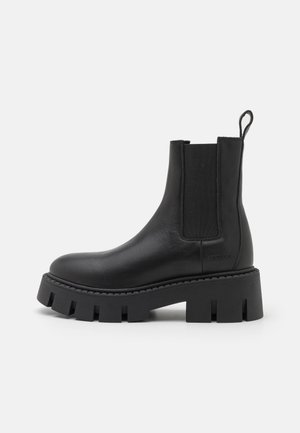 Bota Chelsea de cuero negro con suela gruesa de tacos, paneles laterales elásticos y pestaña para tirar en la parte posterior, mostrada de perfil derecho sobre fondo blanco.