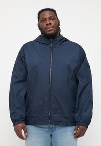 Navy blå hettejakke laget av glatt, lett materiale. Har glidelås, elastiske mansjetter og sidelommer. Enkelt, funksjonelt design.