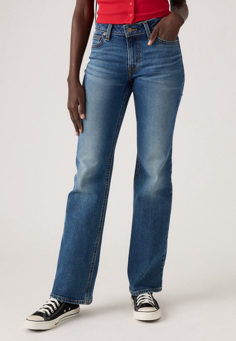 Mannequin portant un jean bootcut bleu délavé moyen, un haut rouge sans manches et des baskets basses noires, debout avec une main dans la poche.