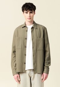 Olivgrön knäppskjorta med två framfickor, texturerad tyg och krage. Buren över en vit t-shirt, matchad med ljusa byxor.