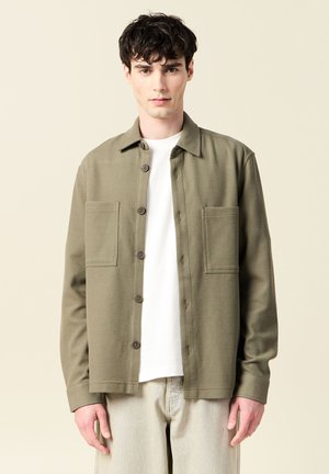 Olive green button-up shirt with two front pockets, textured fabric, and a collar. Worn over a white t-shirt, paired with light-colored pants.
