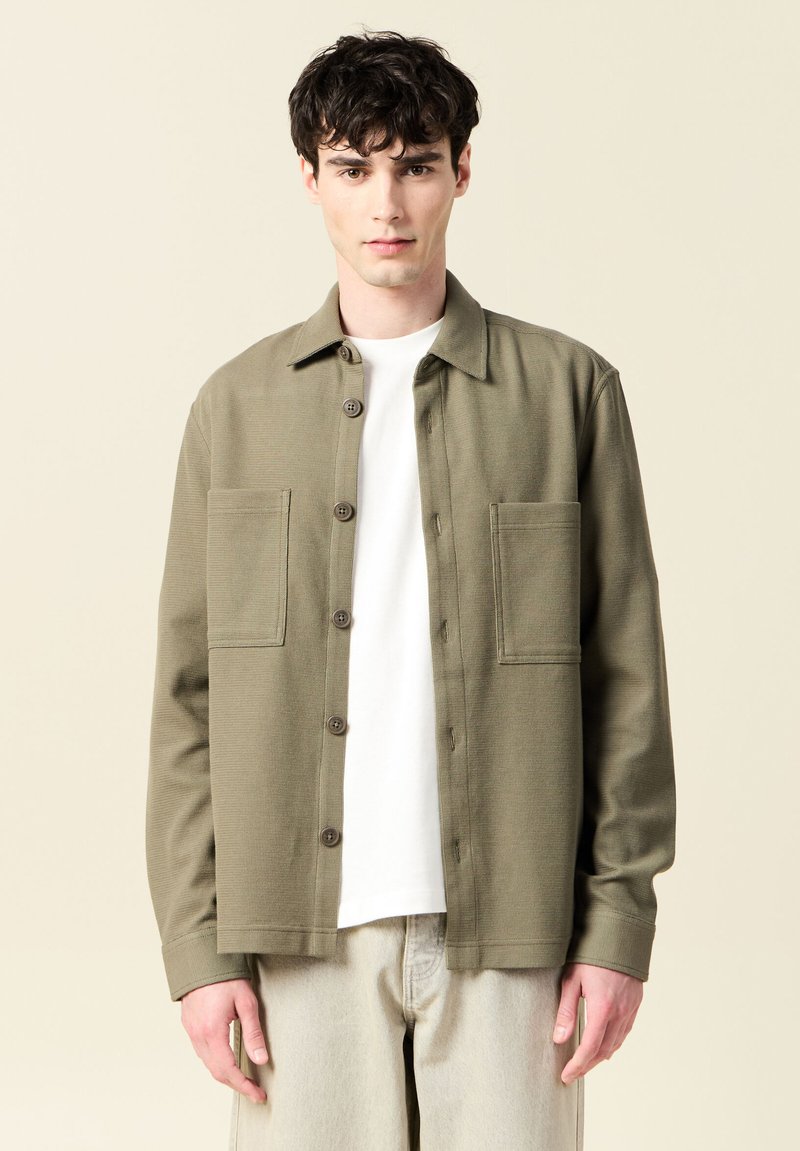 Olivgrön knäppskjorta med två framfickor, texturerad tyg och krage. Buren över en vit t-shirt, matchad med ljusa byxor.