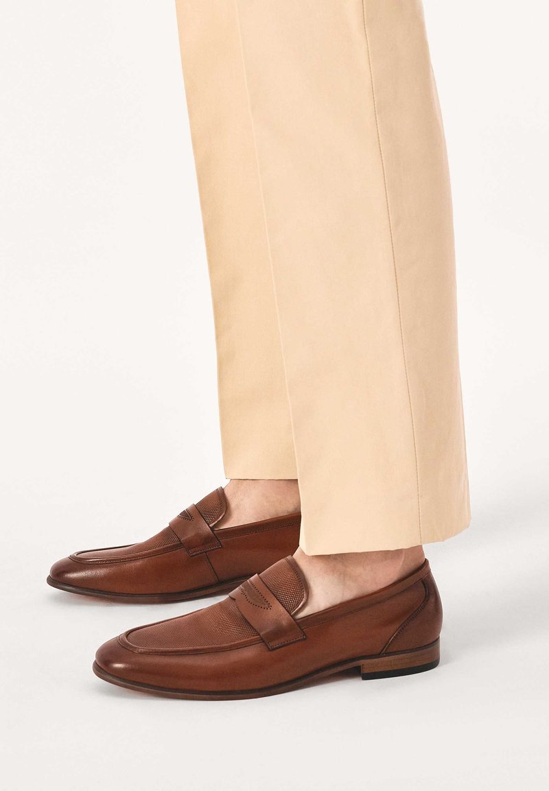 Rudos odinės laisvalaikio batai su lygiu paviršiumi, tekstūruota dirželiu ir žemu kulniuku, derinami su šviesiai smėlio spalvos kelnėmis.