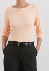 Pfirsichfarbene Langarmbluse mit weitem Ausschnitt, kombiniert mit schwarzen Hosen und einem schwarzen Gürtel mit goldener Schnalle. Weicher Stoff, figurbetonter Schnitt.