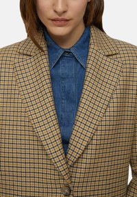 Blazer a quadri giallo e nero, con chiusura a un solo bottone. Indossato sopra una camicia di jeans blu, che mostra texture a contrasto.