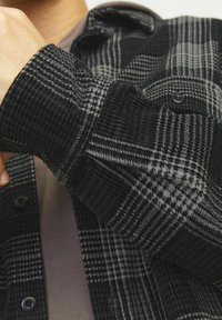 Zwart en grijs geblokte flanellen overhemd met ribbelstructuur, voorzien van een knoopsluiting aan de voorkant en een close-up van de detail van de manchet.