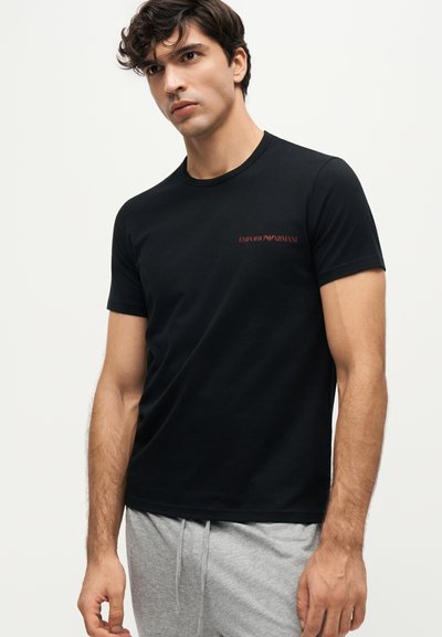 Νέος άνδρας που φοράει μαύρο T-shirt Emporio Armani και ανοιχτό γκρι φόρμας, στέκεται μπροστά σε απλό λευκό φόντο.