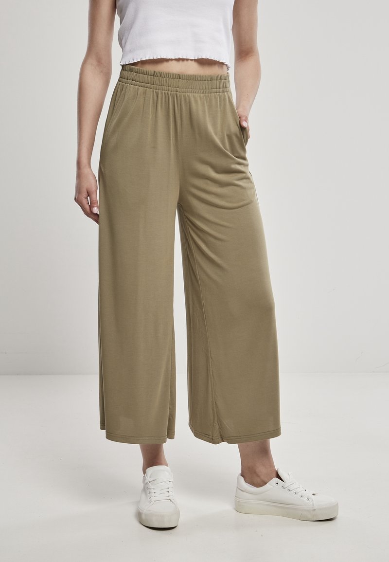 Personne portant un pantalon large ample vert olive, un crop top blanc et des baskets blanches, debout devant un fond clair uni.
