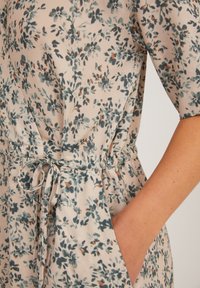 Robe florale beige avec de petites fleurs bleues et oranges, dotée d'une ceinture nouée à la taille et d'une main placée dans une poche latérale.