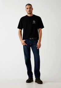 Fekete pamut rövid ujjú póló kis grafikával a mellkasán, kék farmerrel és sötét csizmával párosítva. Egyszerű, hétköznapi kialakítás.