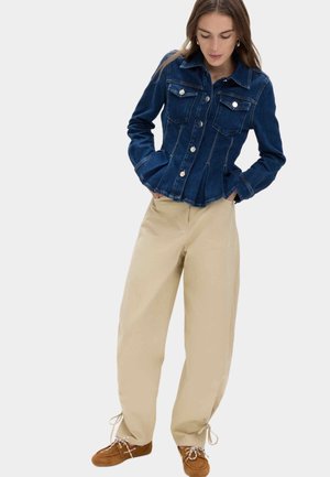 Kvinde iført en mørkeblå denimjakke med sølvknapper, beige bukser med vide ben og brune snørestøvler, stående med hænderne i lommerne.