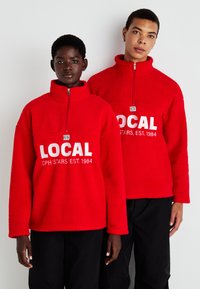 Felpe in pile rosso con collo a mezza zip e testo bianco "LOCAL CPH STARS EST. 1984" sul davanti, indossate da due persone.