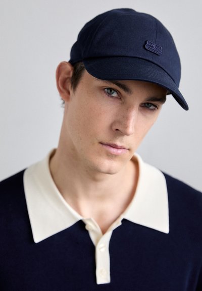 Marinepet met een gebogen rand en een tonale logo. De drager heeft een crèmekleurige polo met een diepe V-hals en knopen. Gelaagd donker gebreid materiaal.