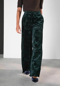 Femeie îmbrăcată cu pantaloni evazați din catifea verde închis, cu un model floral, bluză scurtă maro și pantofi cu vârf ascuțit bleu navy cu fundițe.