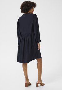 Vestido azul marino con mangas largas, cintura fruncida y una falda fluida. Hecho de una tela ligera, con una superficie sutilmente texturizada.
