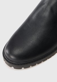 Punta de bota de cuero negro con superficie texturizada y suela de goma gruesa con patrón de profundidad en un fondo blanco.