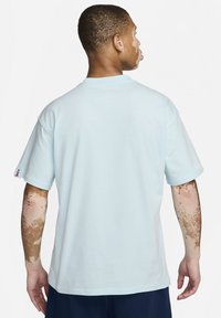 T-shirt en coton bleu clair à manches courtes, coupe ample, col rond côtelé et logo discret sur la manche gauche. Texture lisse.