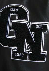 Černá kožená bunda s velkým texturovaným vyšitým písmenem "N" s bílým okrajem a malým vyšitým nápisem "TEAM" a "EST. 1998".