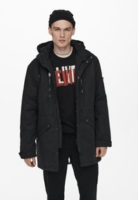 Svart parkas med huva, med dragkedja fram, två sidofickor och justerbara manschetter, bärs över en svart grafisk sweatshirt.