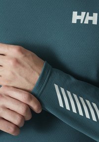 Teal sportieve longsleeve, met textuurstof. Kenmerkt zich door een wit logo en diagonale streepjesdetail op de pols. Close-up van de handpositie op de stof.
