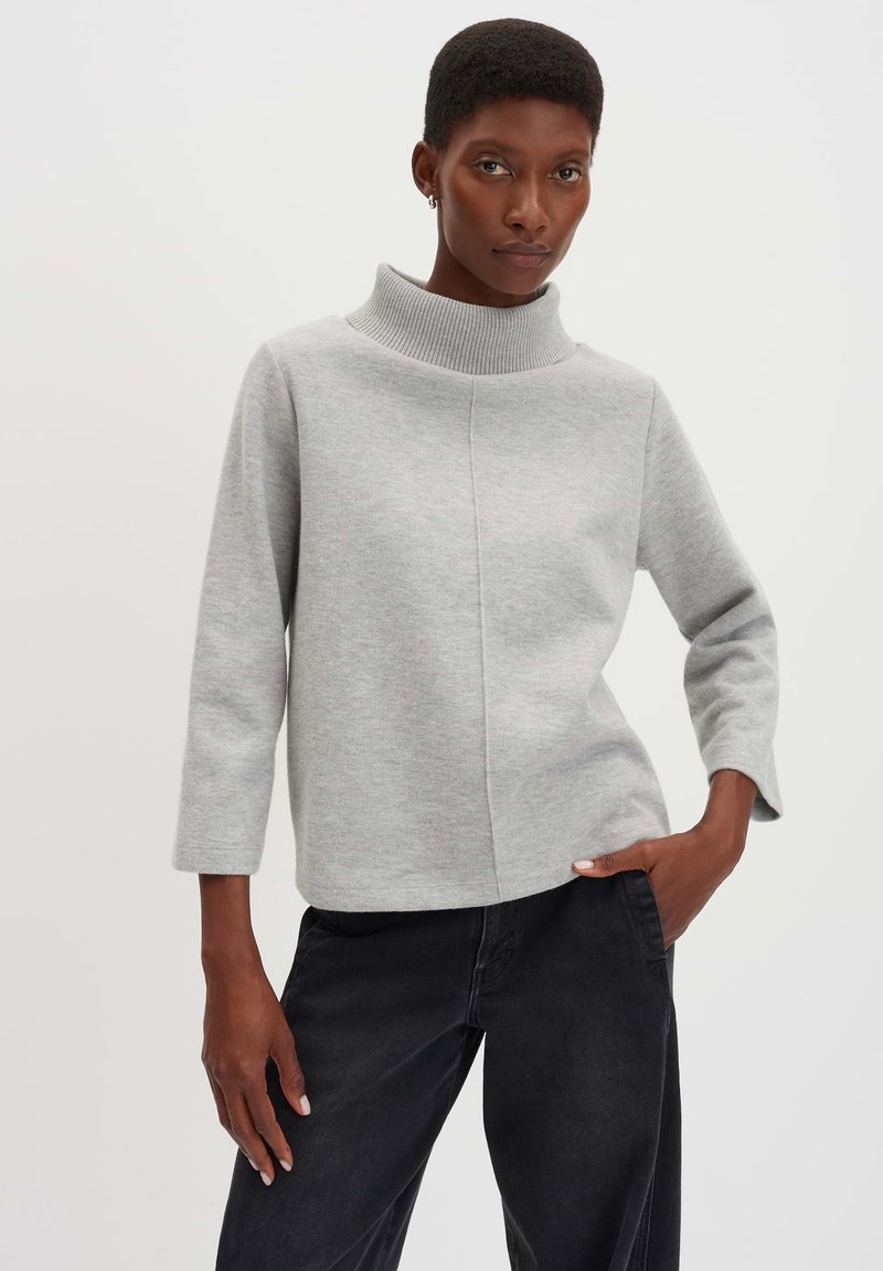 Hellgrauer Pullover mit geripptem Rollkragen, legerem Schnitt, abgesetzten Nähten und Dreiviertelärmeln. Trägt man zu schwarzen Hosen.